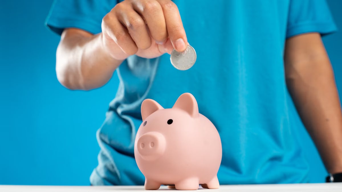 A close-up of an adult's hand dropping a coin into a piggy bank, symbolizing savings and investment.